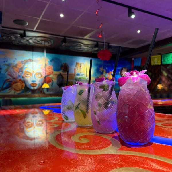 Four colorful El Leoncito cocktails in pineapple-shaped glasses on a red bar with a mural.