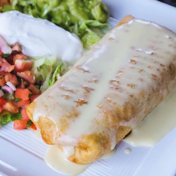 A golden-brown chimichanga covered in white cheese sauce, served with guacamole, sour cream, and pico de gallo.