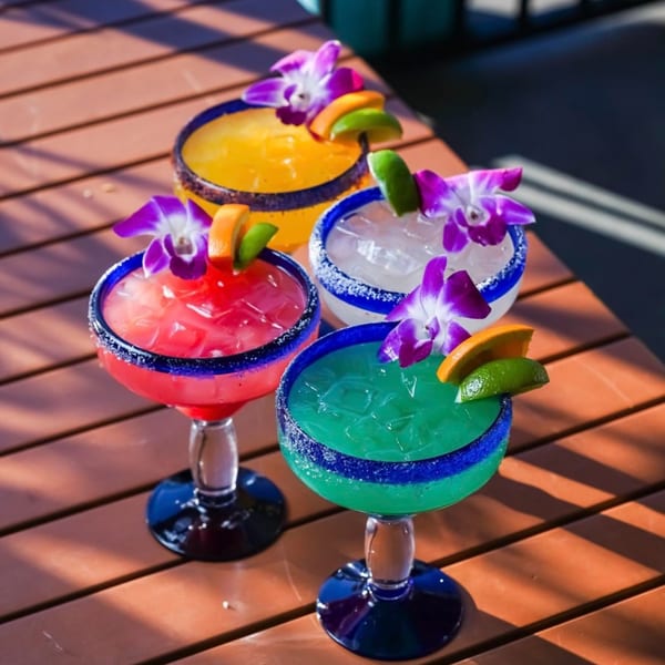 Four colorful margaritas with fruit garnishes and purple orchids on a wooden table.
