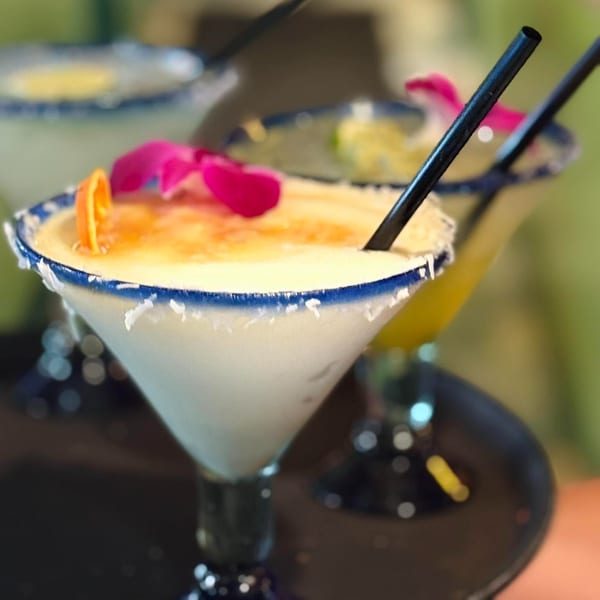 Close-up of a creamy margarita with an orange slice and pink flower, rimmed with salt.