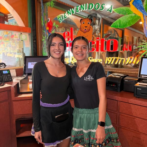 Two smiling El Leoncito restaurant staff members stand at the reception desk with colorful decor.