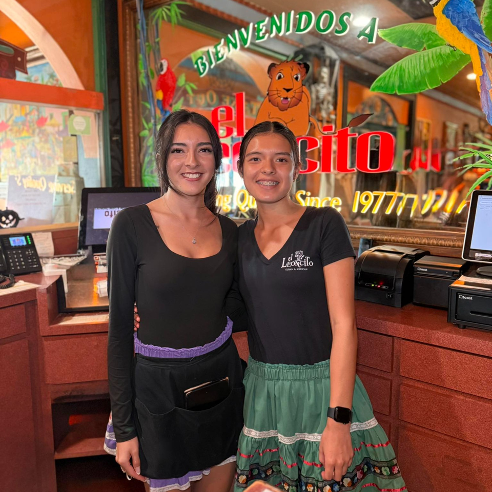 Two smiling El Leoncito restaurant staff members stand at the reception desk with colorful decor.