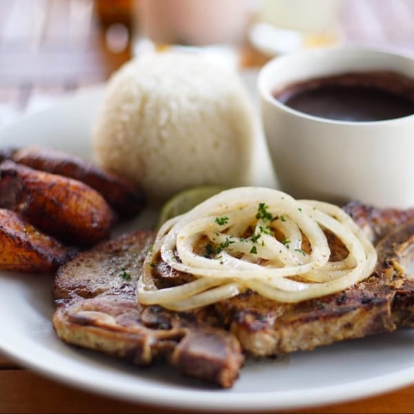 Grilled pork chop topped with onions and parsley, served with rice, black beans, and plantains.