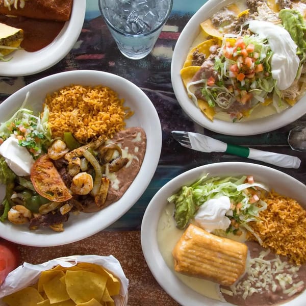 Overhead view of four plates of Mexican food, including fajitas, nachos, and enchiladas, with chips and water.