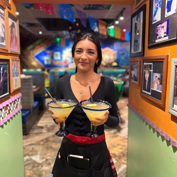 A smiling El Leoncito server holds two margaritas in a vibrant, decorated Mexican restaurant.