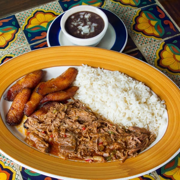 A plate of shredded beef, white rice, fried plantains, and a side of black beans with onions.