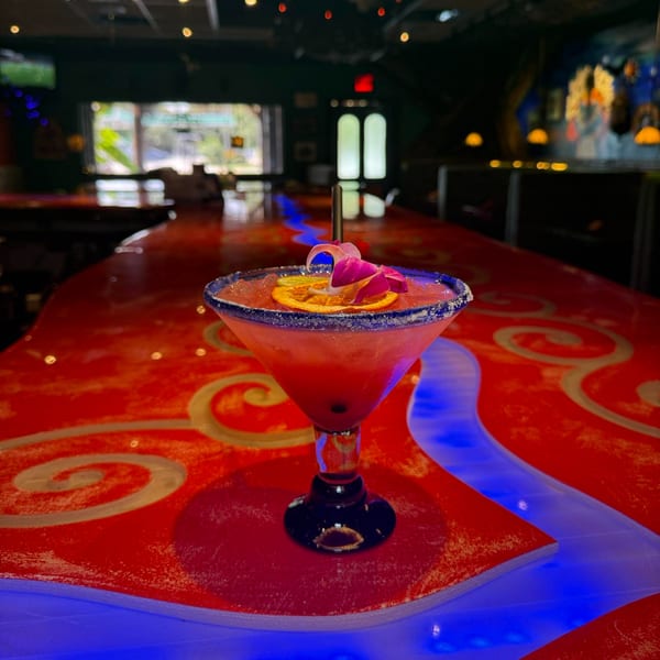 A vibrant pink margarita with orange and purple flower garnish sits on a red bar with blue accents.